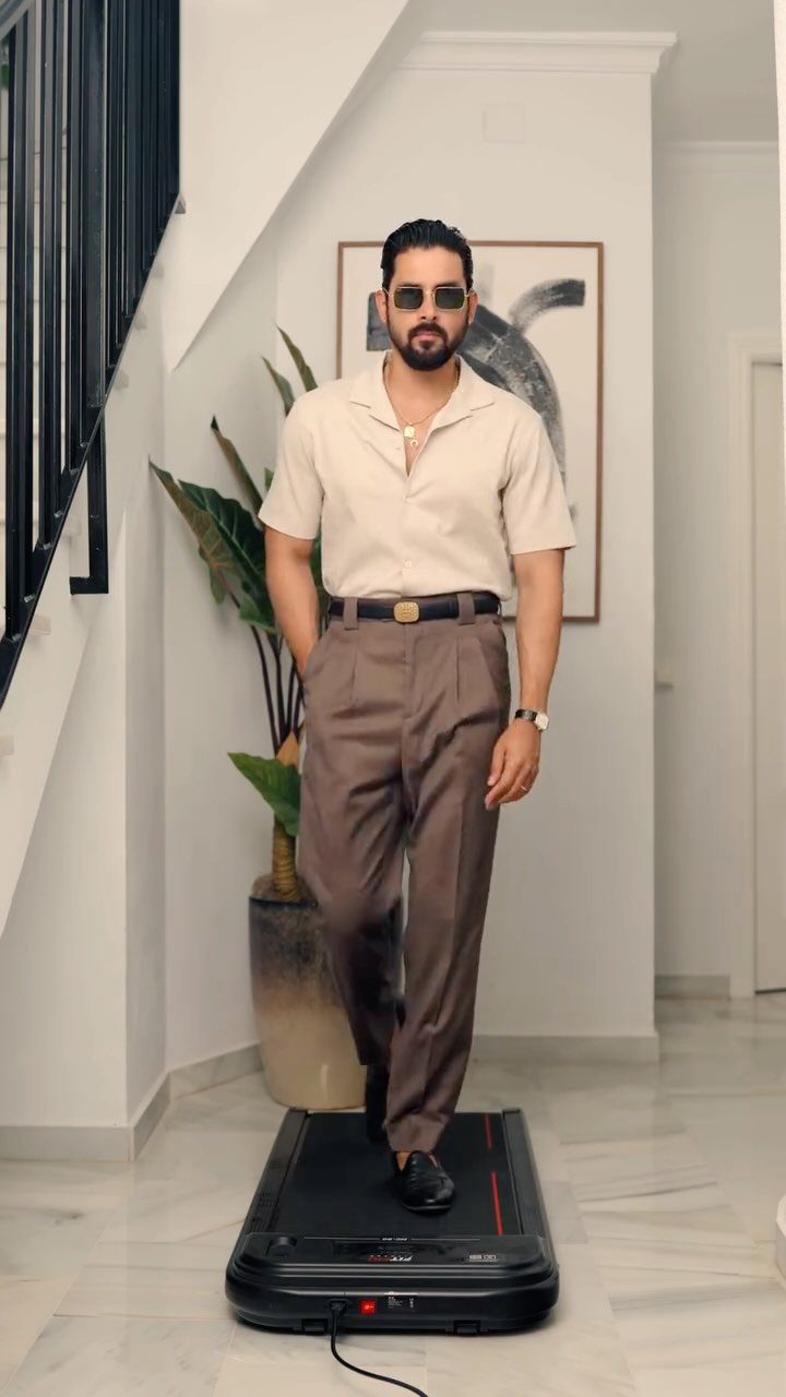 A man in a cream shirt and brown trousers walking on a treadmill indoors.