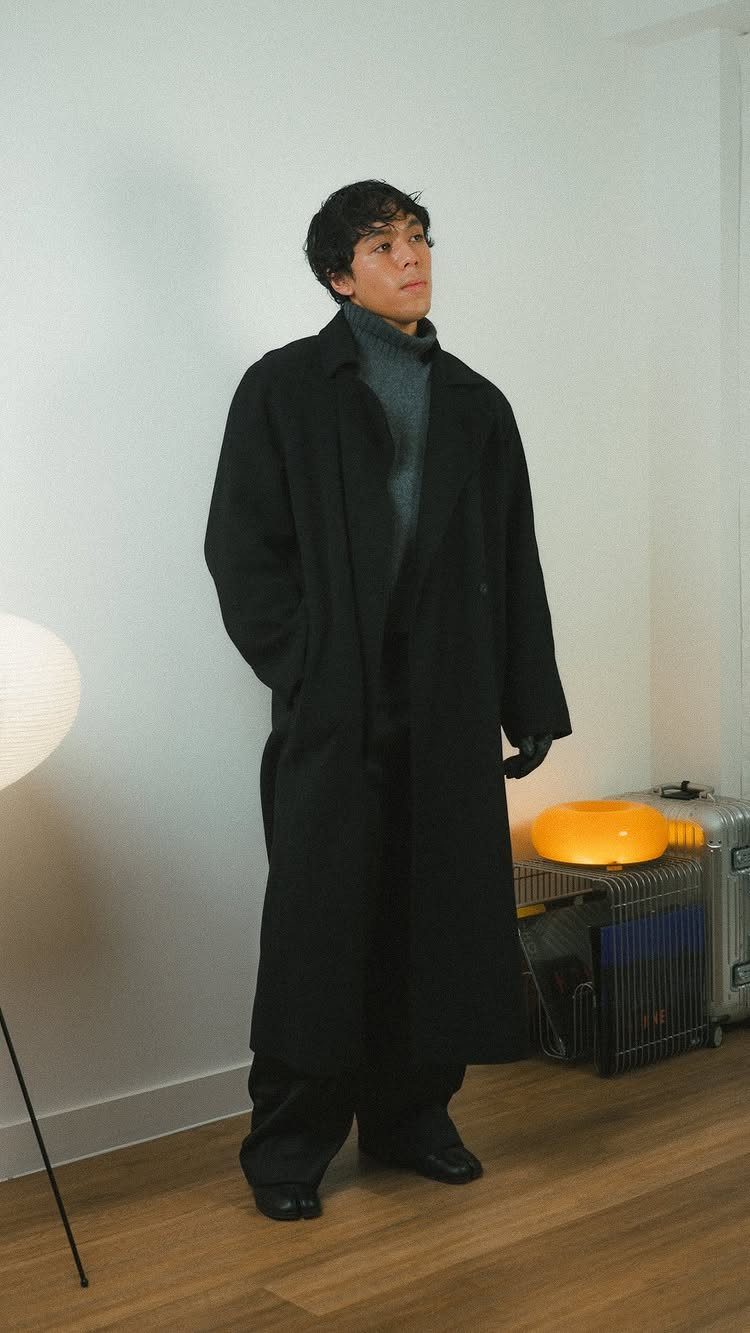 A man wearing a long black overcoat layered over a grey turtleneck sweater and dark trousers.