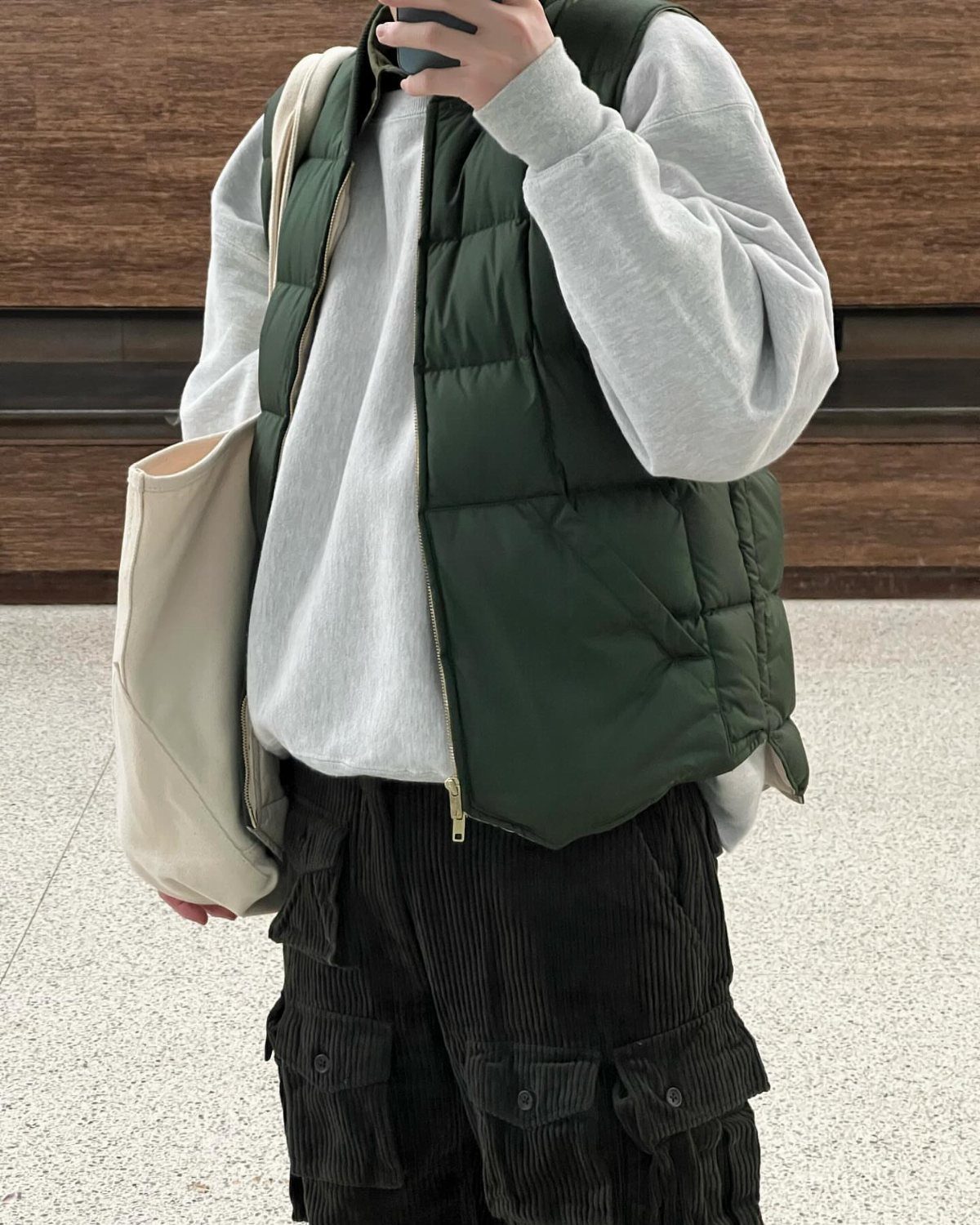 Person wearing a grey sweatshirt, green puffer vest, and dark corduroy cargo pants with a tote bag.