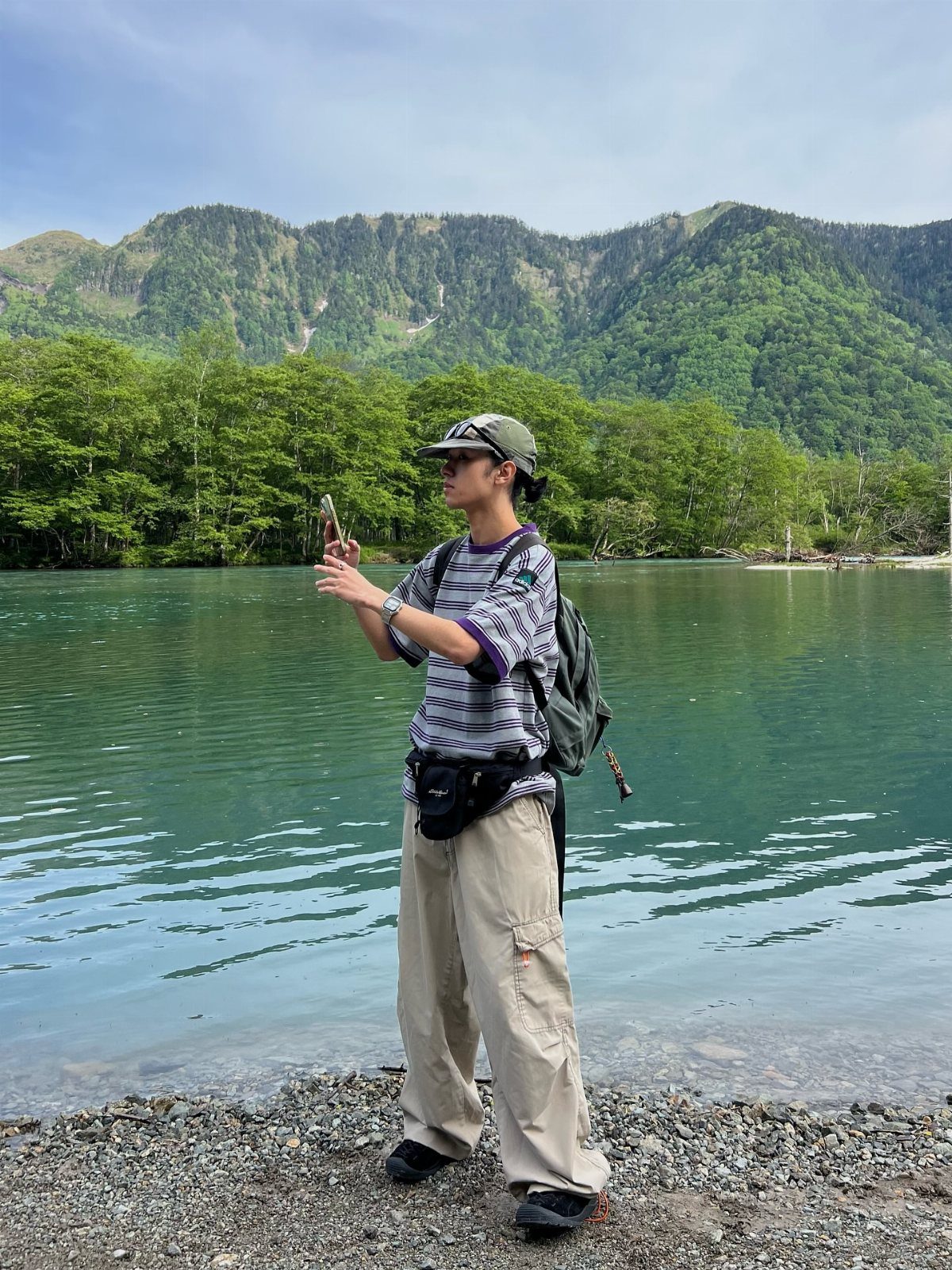 A young man in outdoor clothing stands by a turquoise lake with mountains in the background.