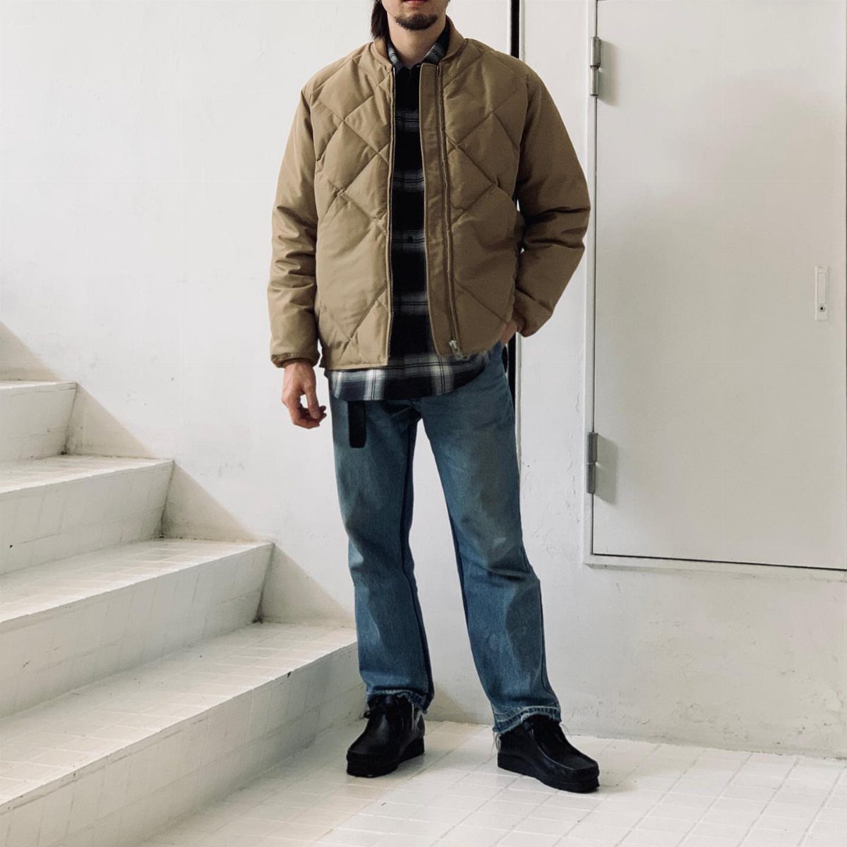 Man wearing a tan quilted jacket, plaid shirt, and blue jeans standing by white stairs.