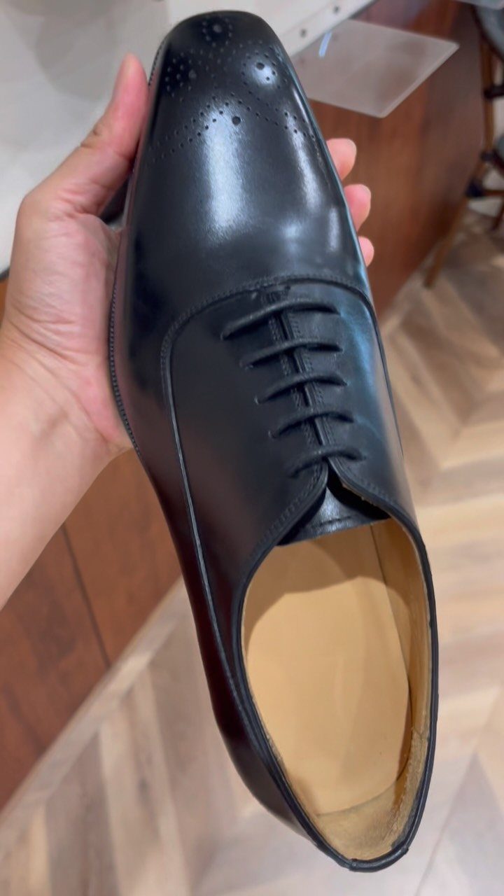 A hand holding a polished black leather lace-up dress shoe with decorative toe perforations.