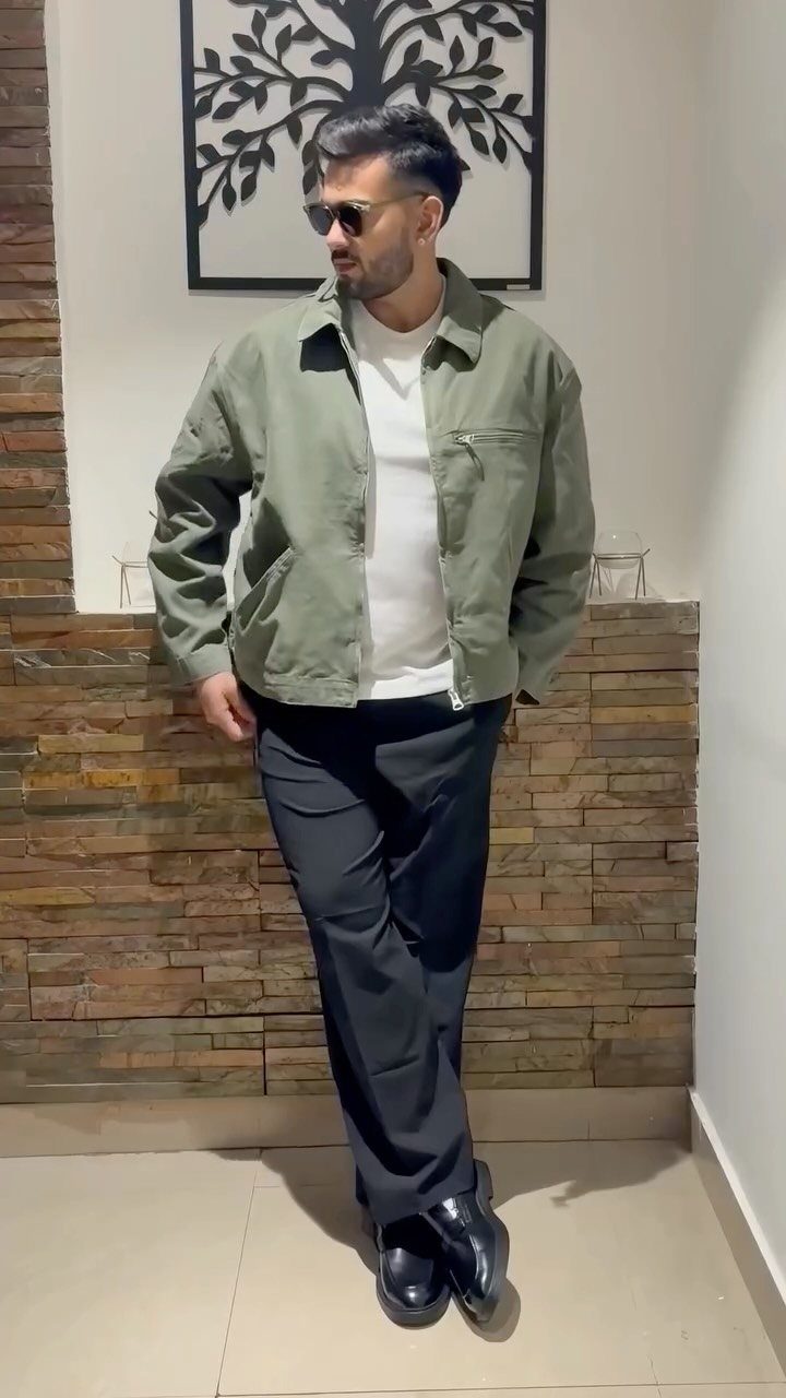 Man in an olive green jacket, white t-shirt, black trousers, and black shoes standing indoors.