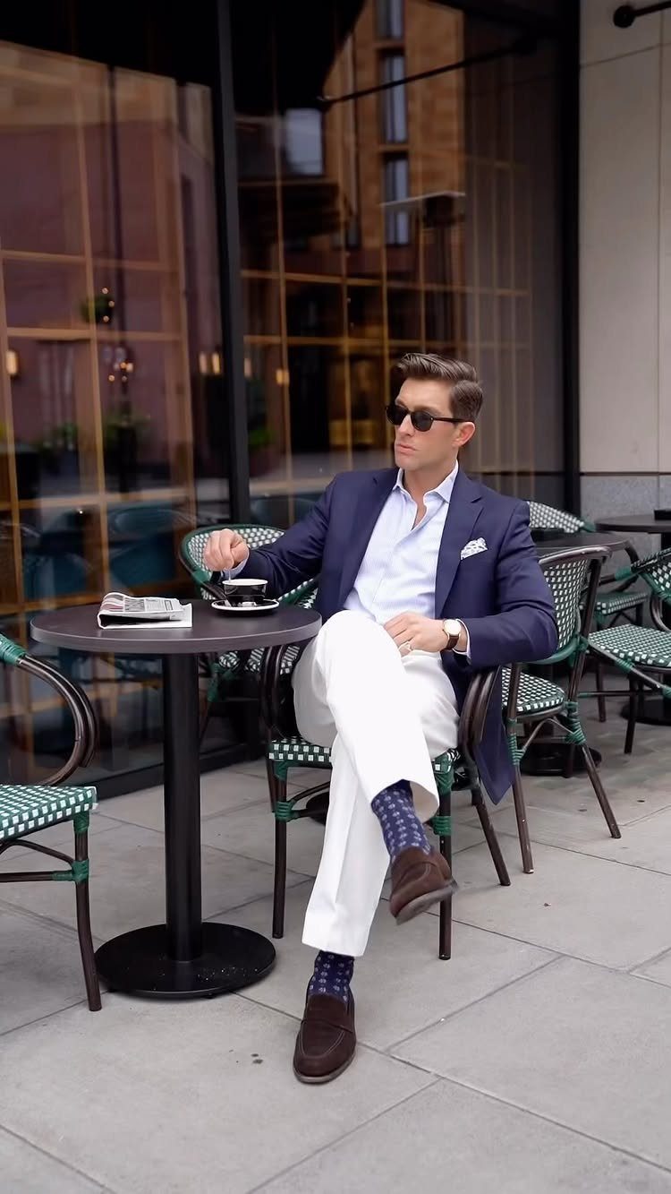 Man in a navy blazer and white trousers sitting at an outdoor cafe table.