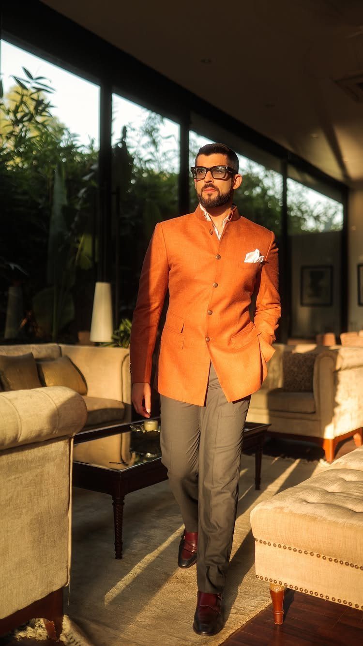 Man in an orange Nehru jacket and grey trousers standing in a sunlit living room.