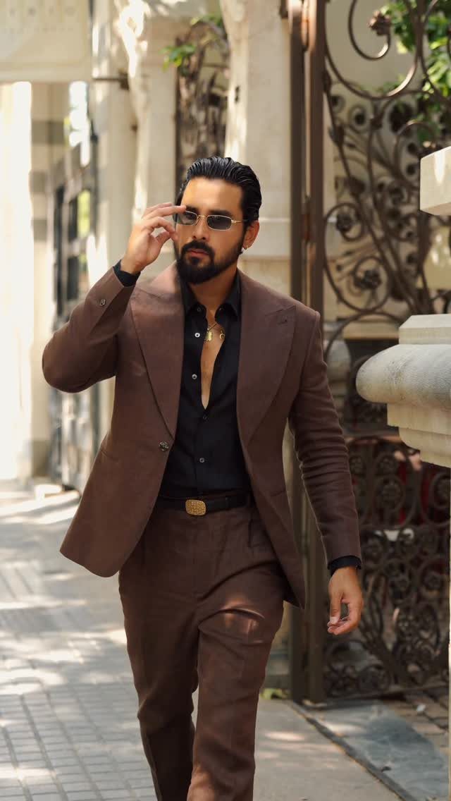 A man walking outdoors wearing a perfectly tailored brown suit and black dress shirt.