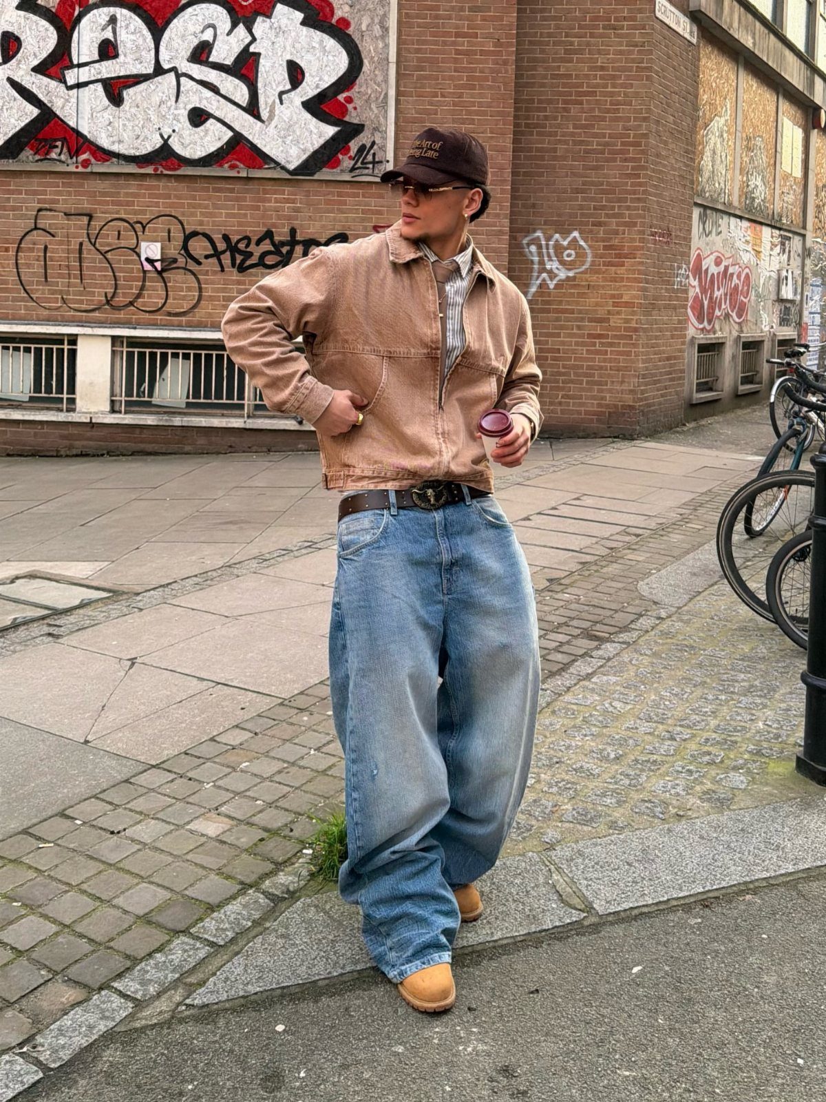 Man wearing a tan boxy jacket and baggy light-wash blue jeans standing on a city street.