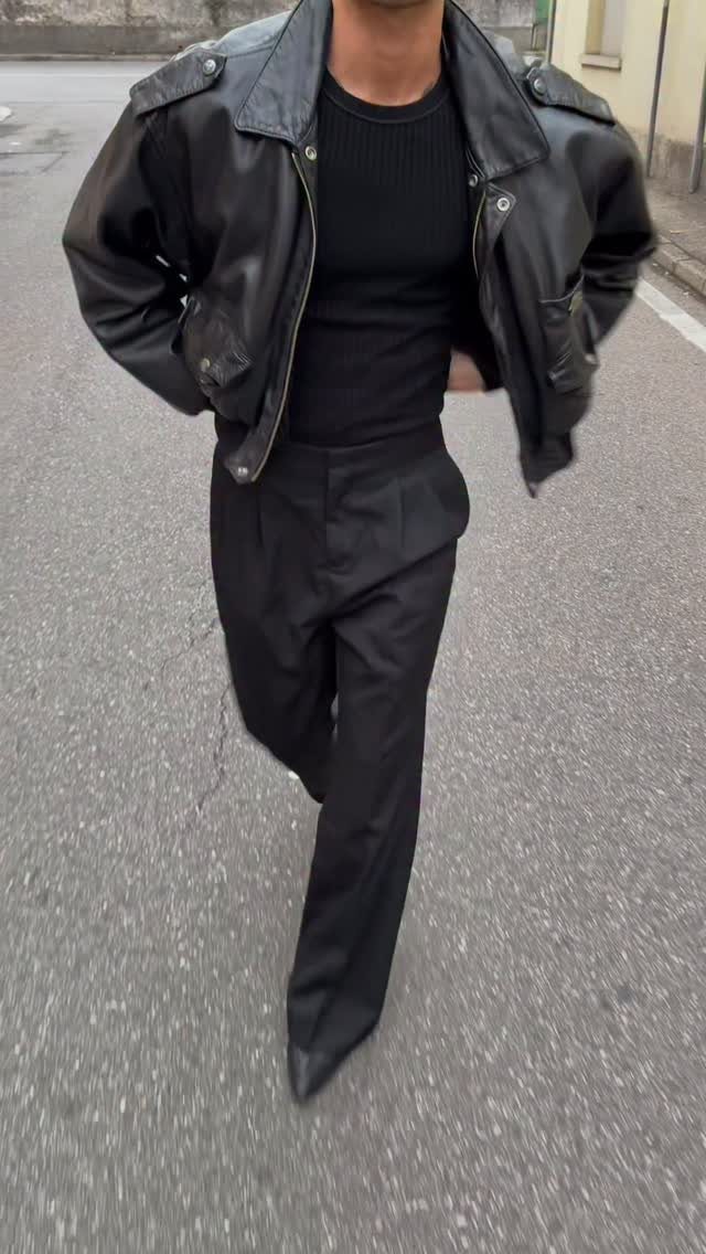 Man walking on an asphalt street wearing an oversized black leather jacket and black trousers.