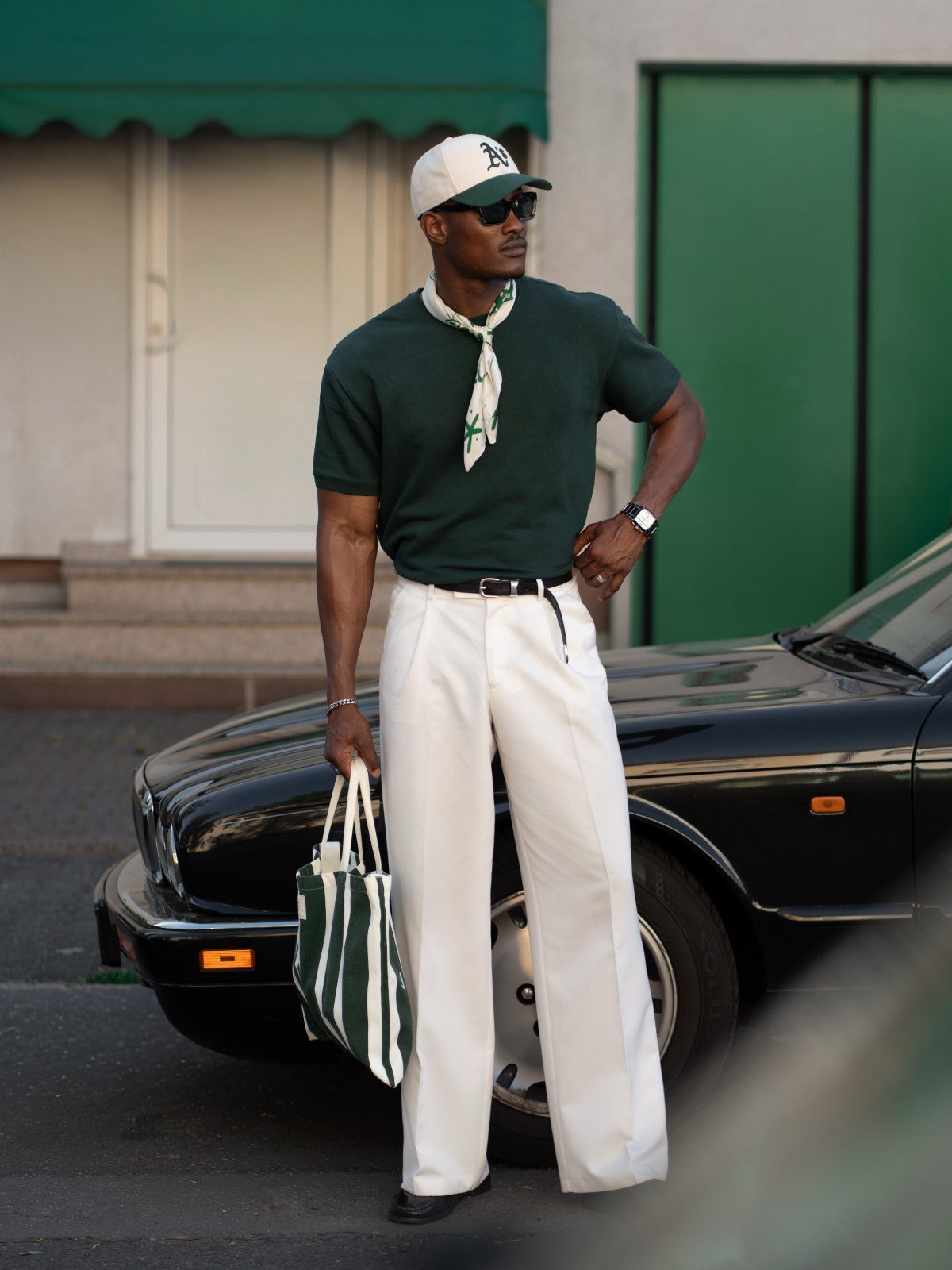 Man in dark green t-shirt and white wide-leg trousers standing by a black car.