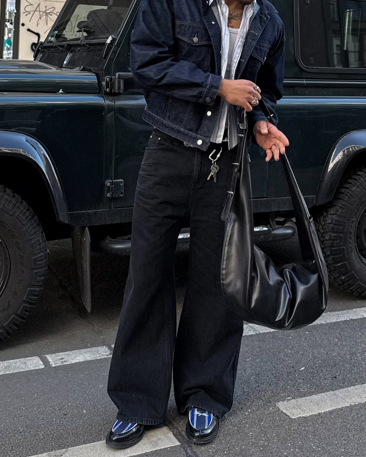 Man wearing a dark denim jacket, white shirt, wide-leg black trousers, and black loafers standing by a vehicle.