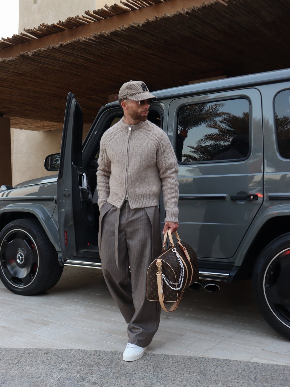 Man in elevated streetwear standing next to a grey luxury SUV carrying a designer bag.