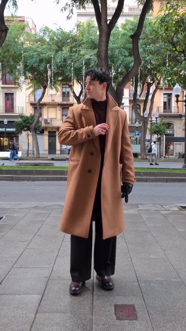 A man wearing a long camel coat and black wide-leg trousers standing on a city street.