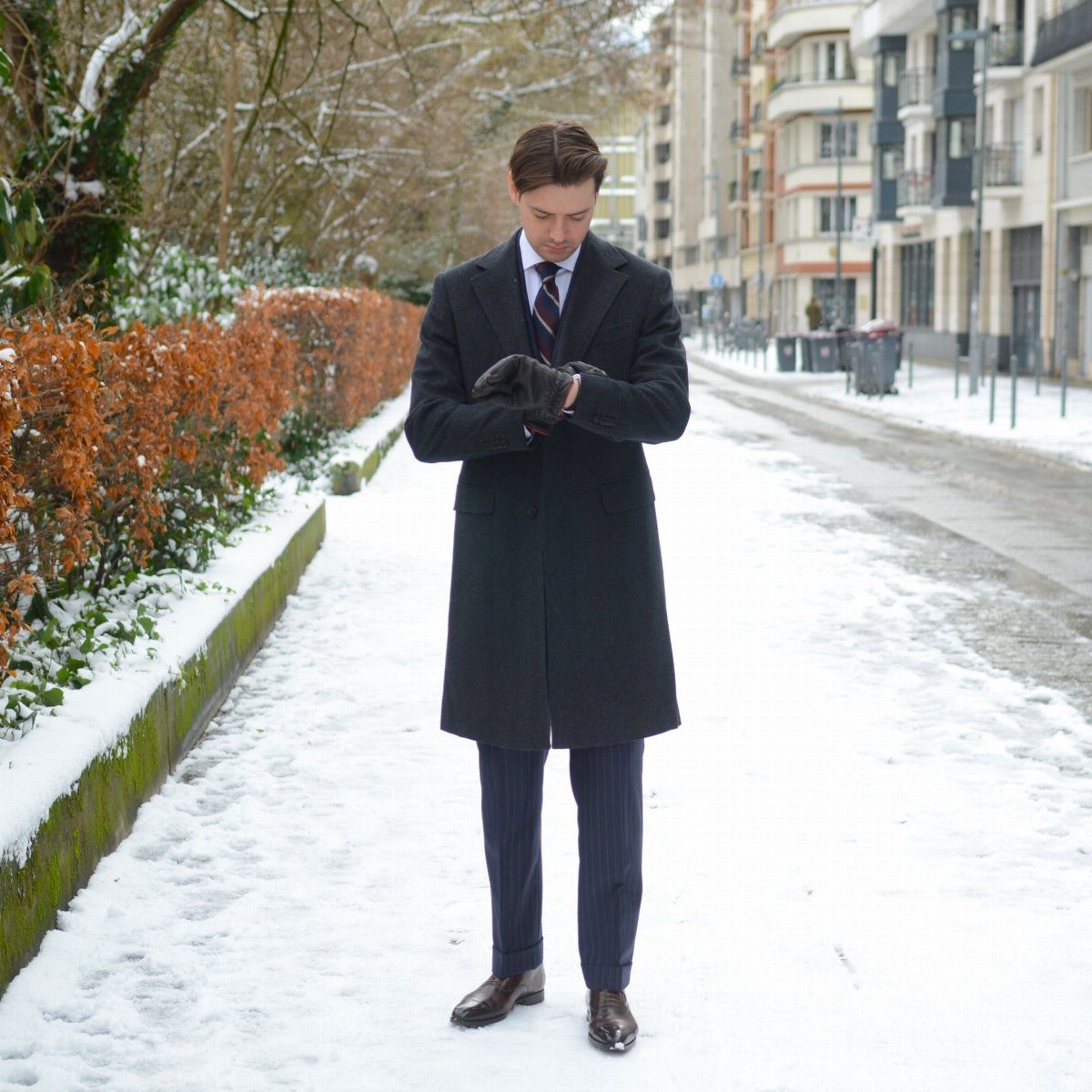 A man in a long black coat and pinstriped trousers standing on a snowy sidewalk.