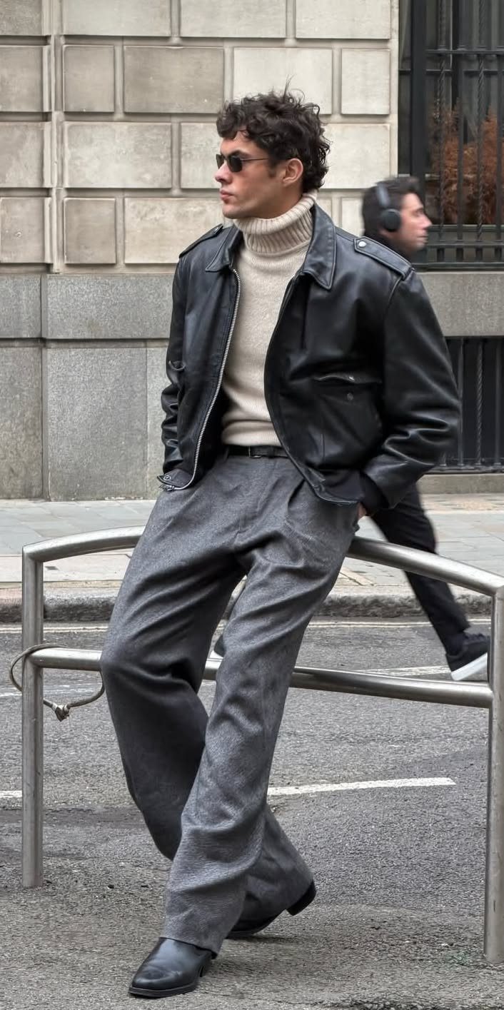 Man in a cream turtleneck, black leather jacket, and grey trousers leaning against a metal railing.