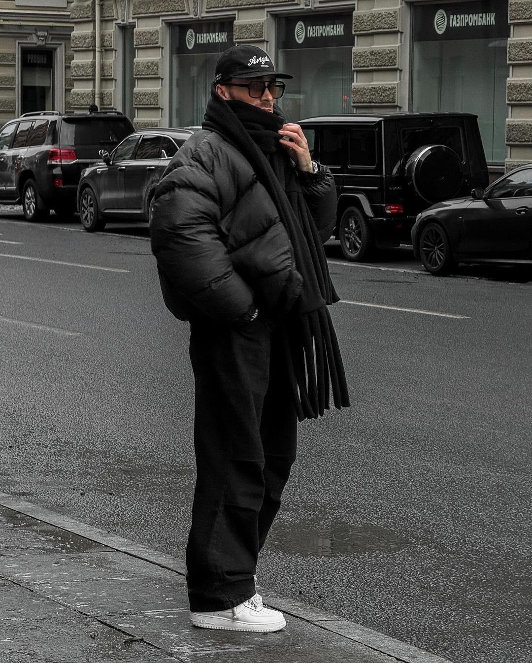 A person wearing a black puffer coat, black trousers, and a black scarf standing on a city street.