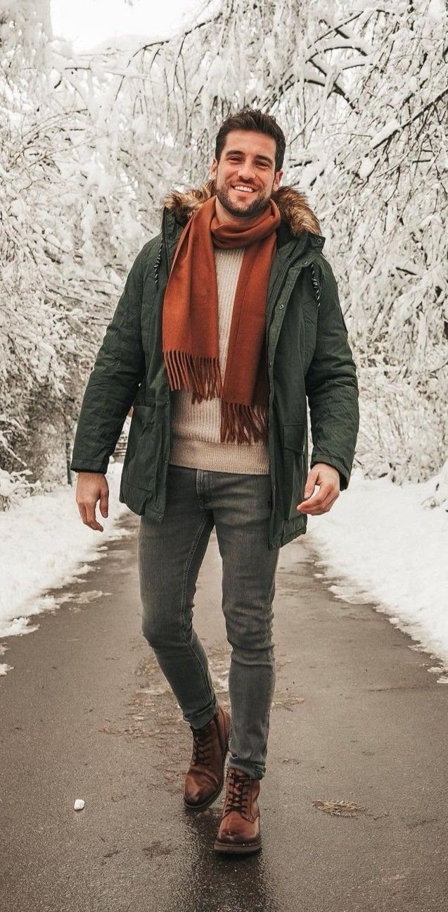 A smiling man in a green parka, orange scarf, and grey jeans walking through a snowy forest.
