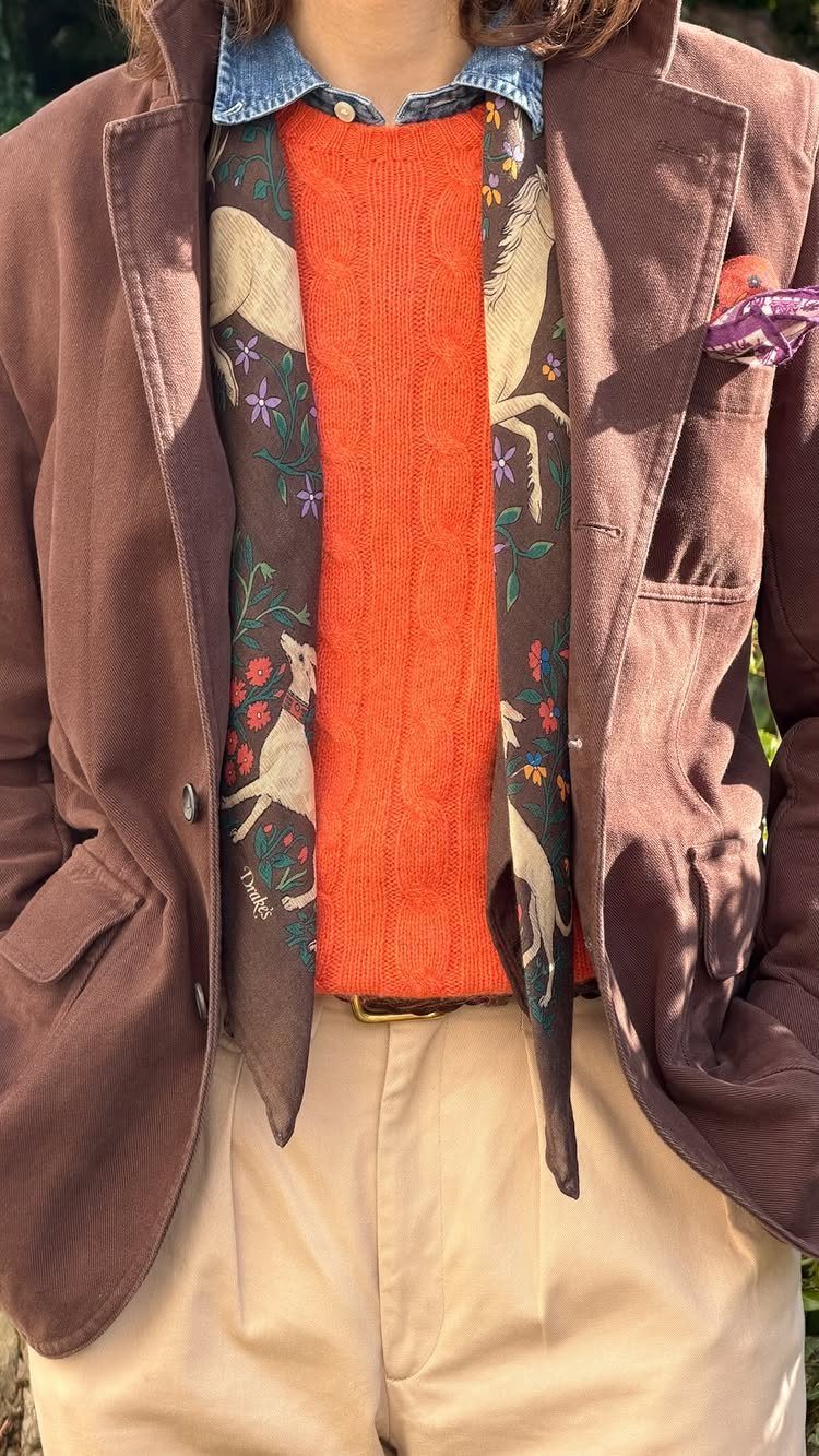 A person wearing an orange knit top, brown blazer, patterned scarf, and beige trousers.