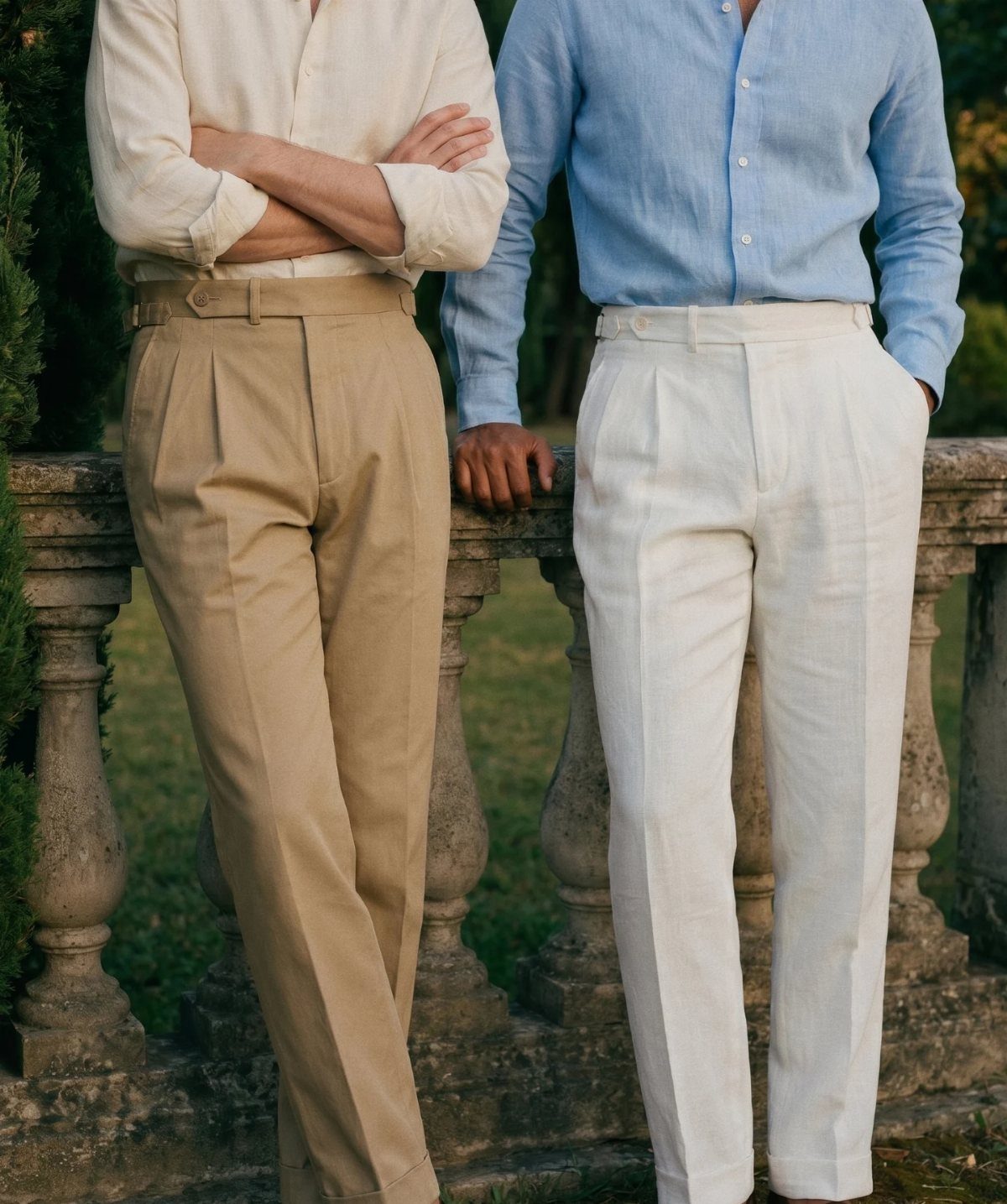 Two men wearing pleated linen trousers and button-down shirts standing by a stone railing.