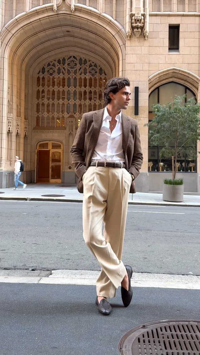 Man in a brown blazer and beige trousers posing on a city street near an arched building.