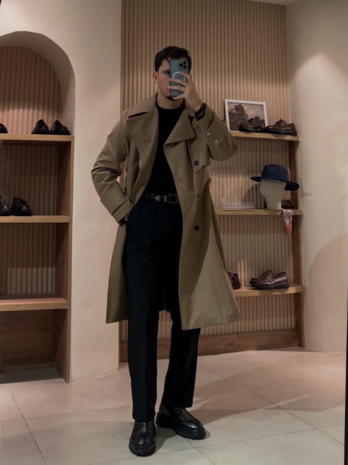 Man in a tan trench coat, black turtleneck, and black trousers posing for a mirror selfie.