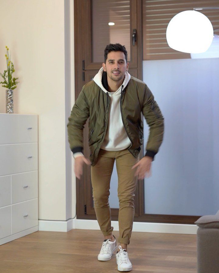 Man walking indoors wearing an olive bomber jacket, white hoodie, tan pants, and white sneakers.