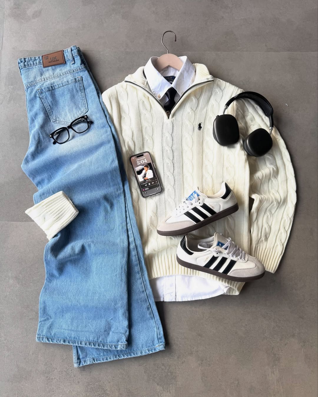 Flat lay of light blue jeans, white cable-knit sweater, white sneakers, and black headphones on grey background.