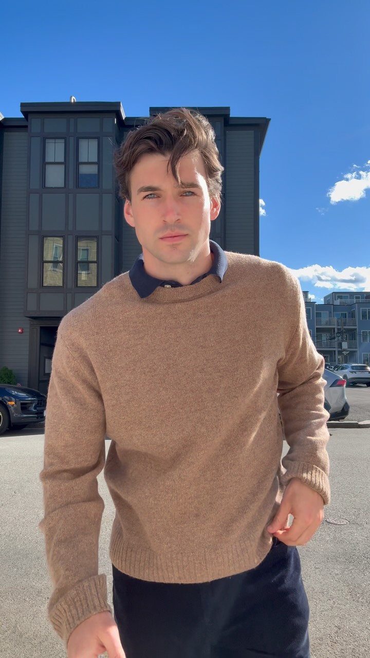 A young man wearing a brown knit sweater and dark collared shirt standing outdoors.