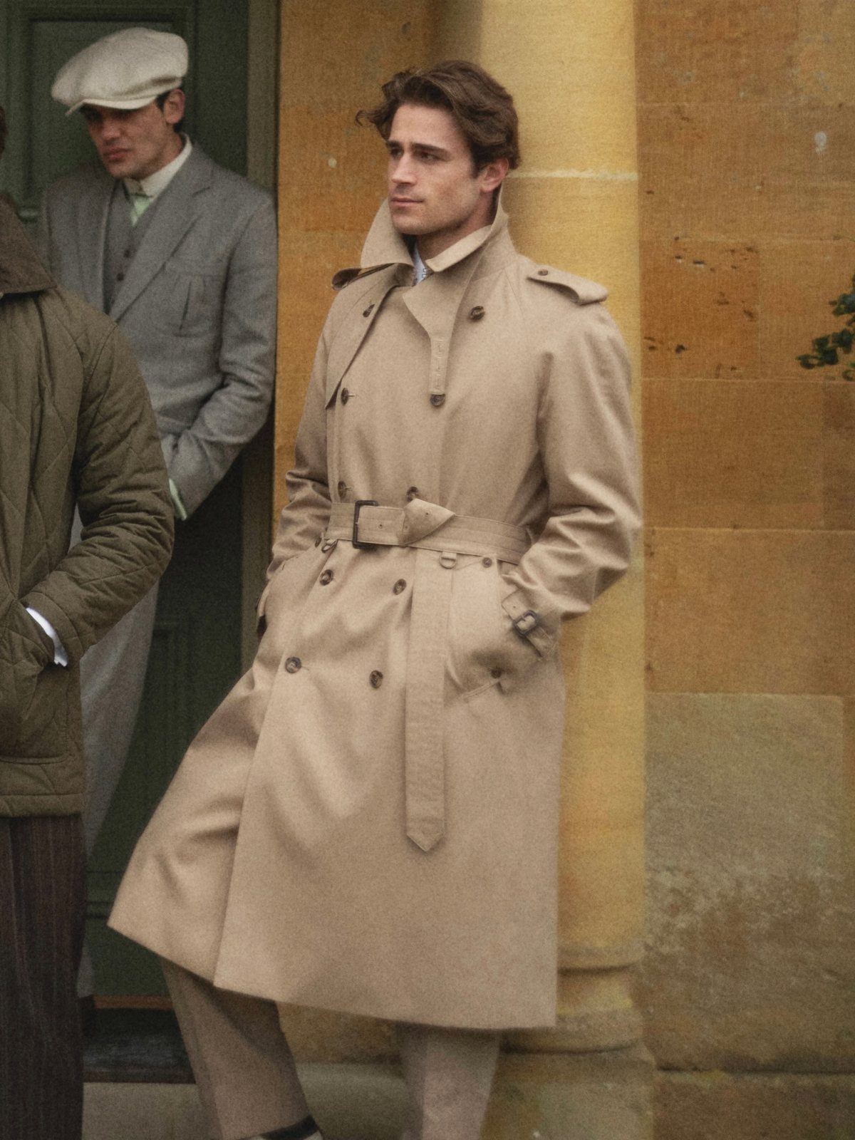 A man wearing a long tan belted trench coat standing against a stone wall.