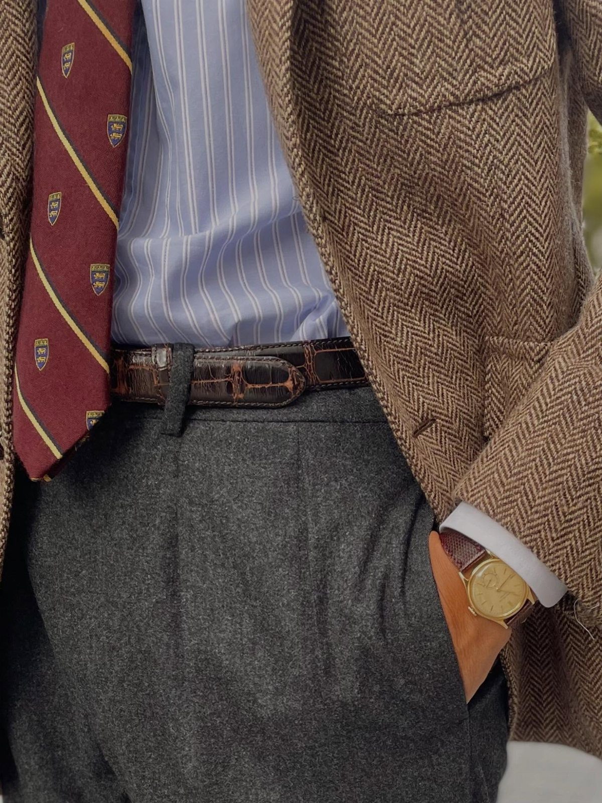 A person dressed in a brown herringbone blazer, striped shirt, maroon tie, and grey trousers.