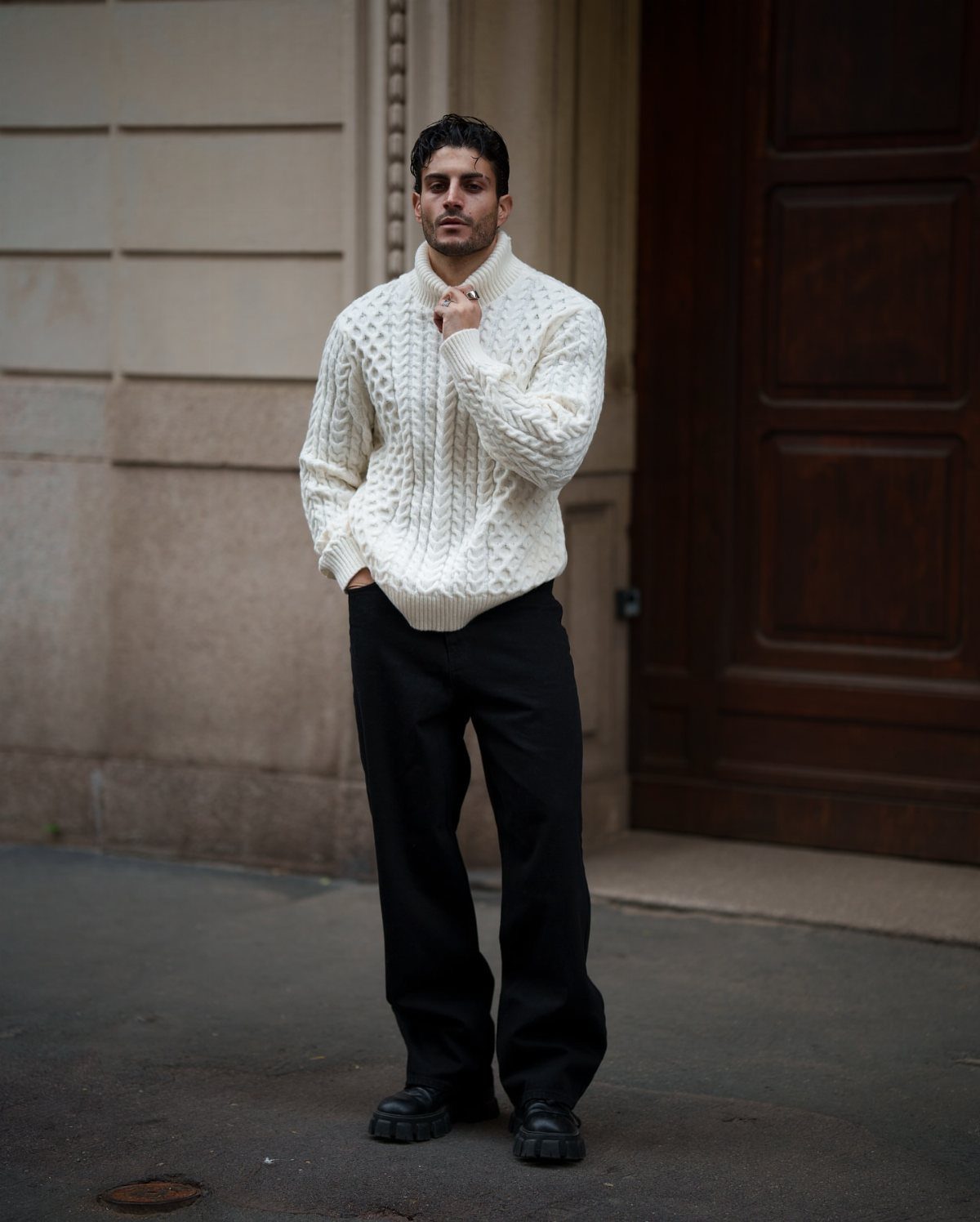 A man wearing a white cable-knit turtleneck sweater and wide-leg black trousers standing outdoors.