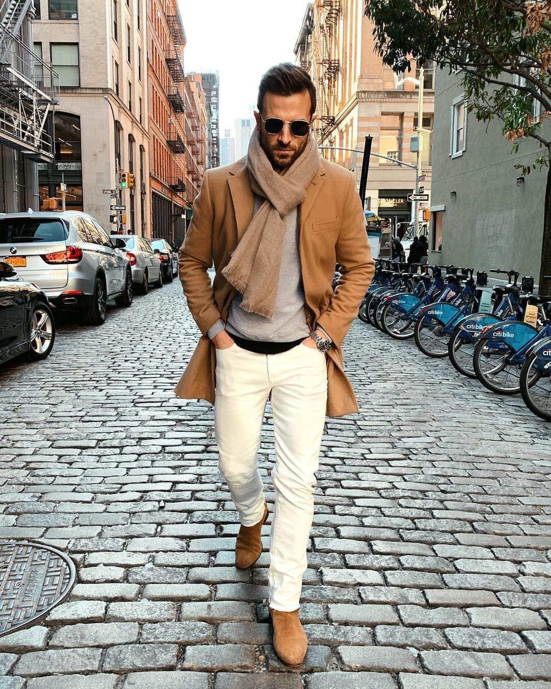 Man in a tan coat, beige scarf, and slim white jeans walking on a cobblestone street.