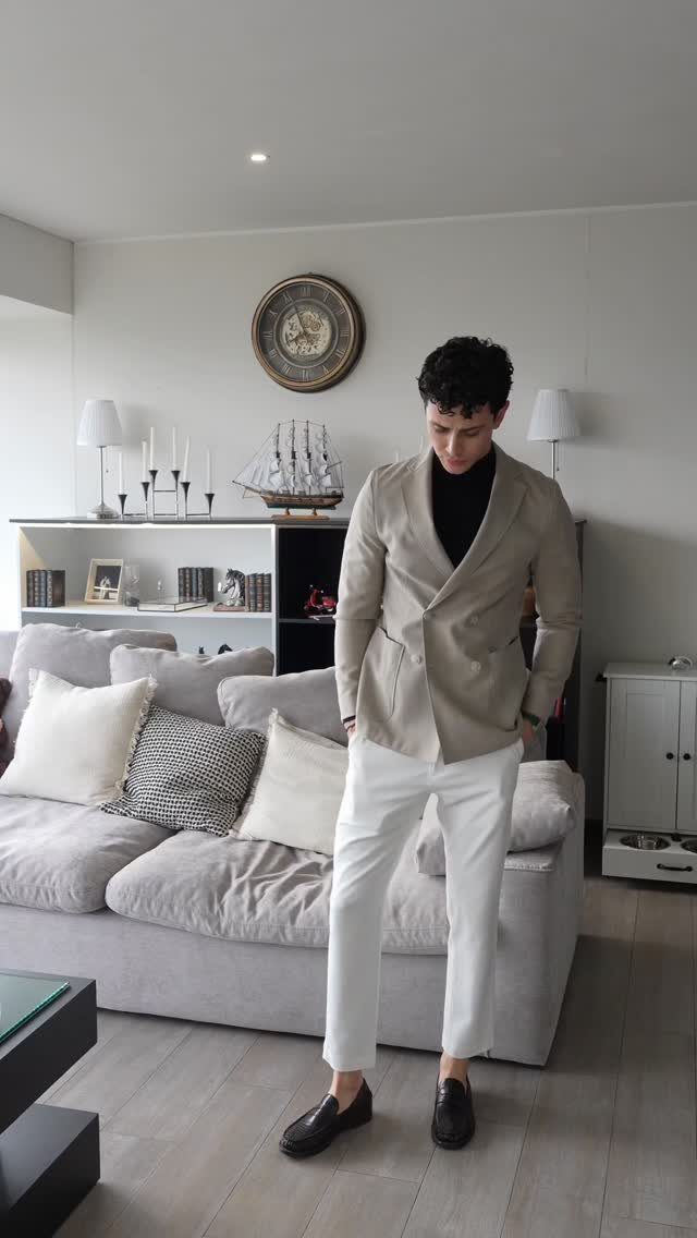 A man wearing a beige blazer, black turtleneck, and white trousers standing in a living room.