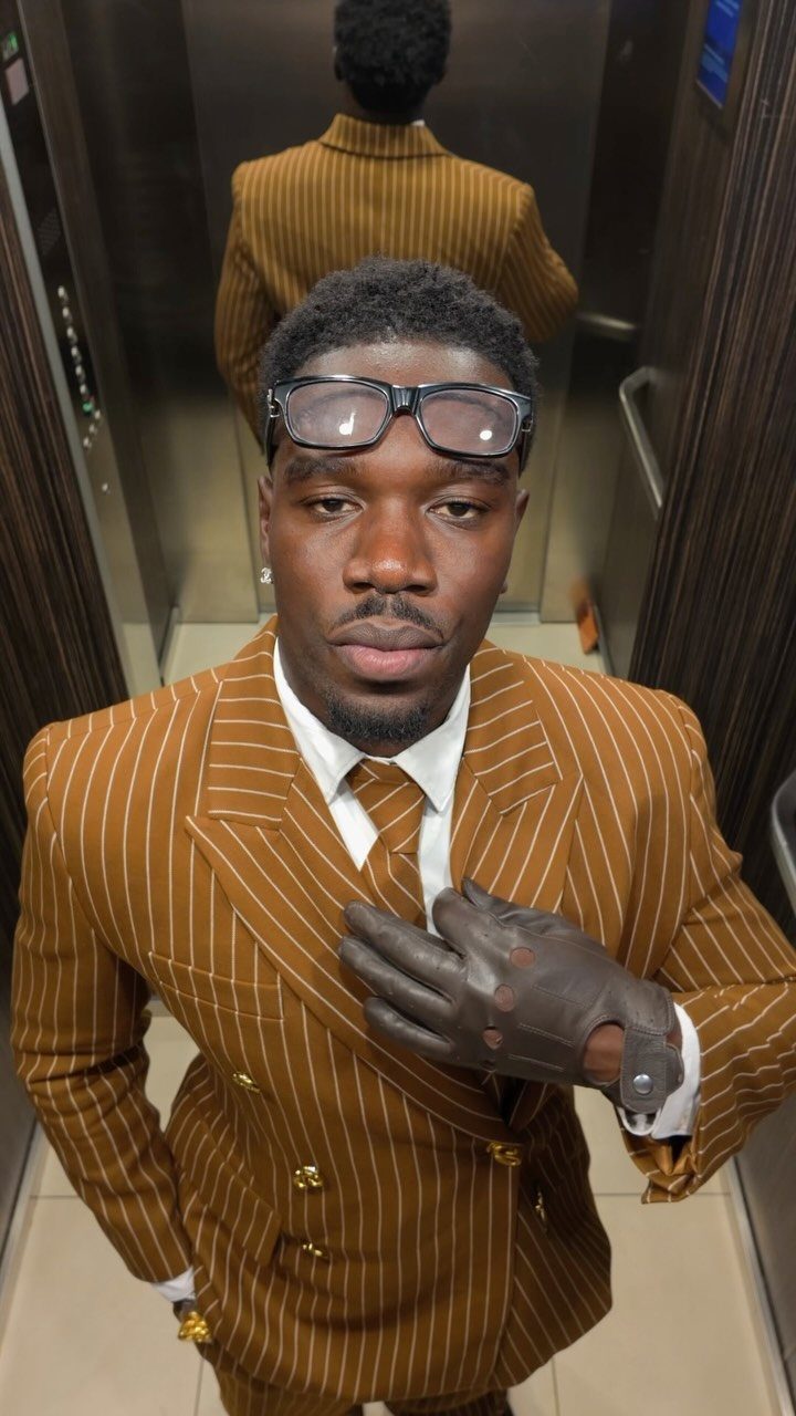 A man wearing a brown pinstripe suit and leather gloves posing in an elevator.