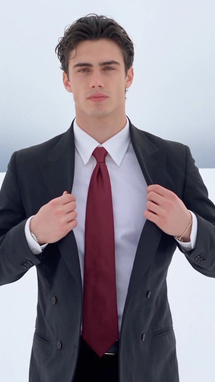 A young man wearing a charcoal grey suit, white shirt, and burgundy tie against white.