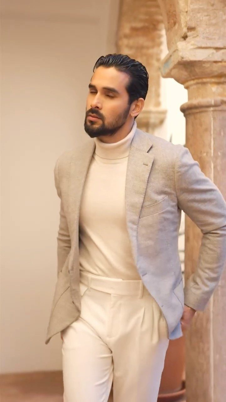 A man wearing a grey blazer, cream turtleneck, and white trousers standing indoors.