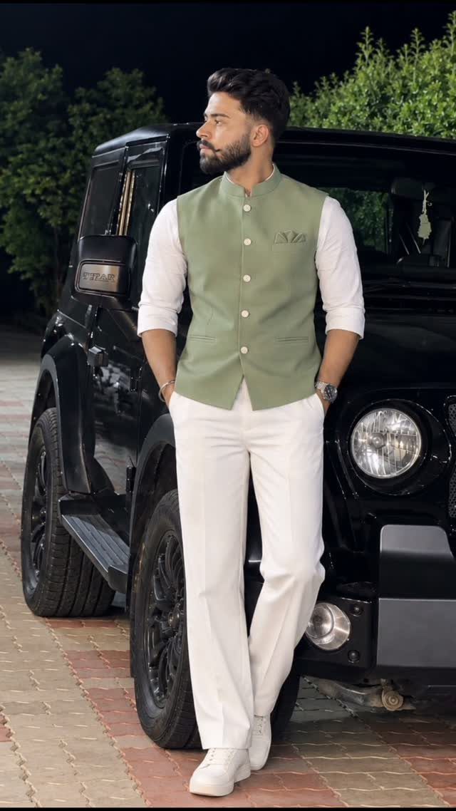 A man posing in a green vest and white trousers leaning against a black SUV.