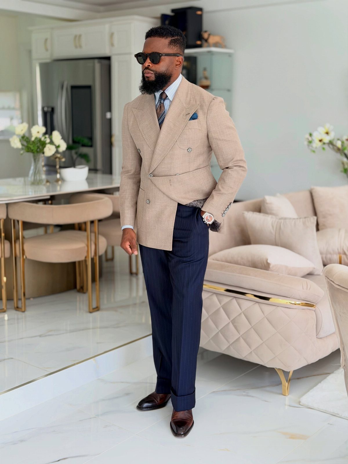 Man in a tan double-breasted blazer and navy pinstripe trousers standing in a modern room.