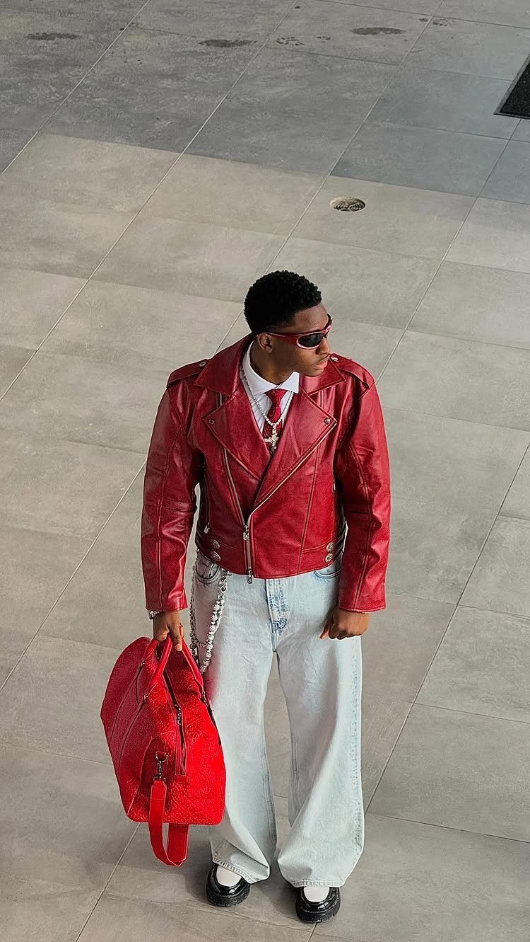 A man in a red leather jacket, white shirt, red tie, and light blue jeans standing outdoors.