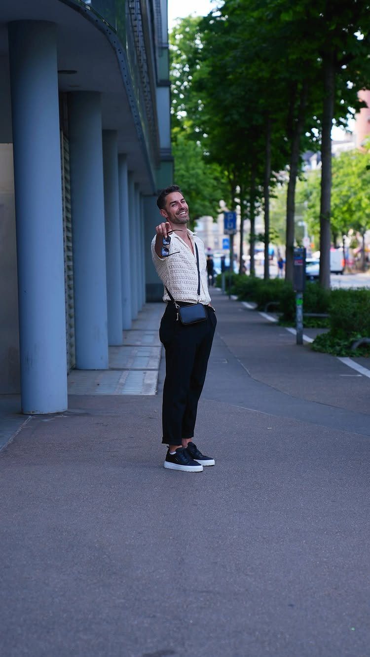 A smiling man in a patterned cream shirt and black trousers posing on a city sidewalk.