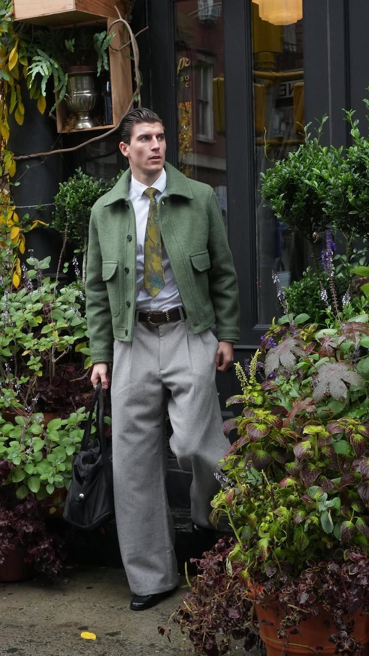 A man dressed in a green jacket, white shirt, tie, and grey trousers standing among plants.