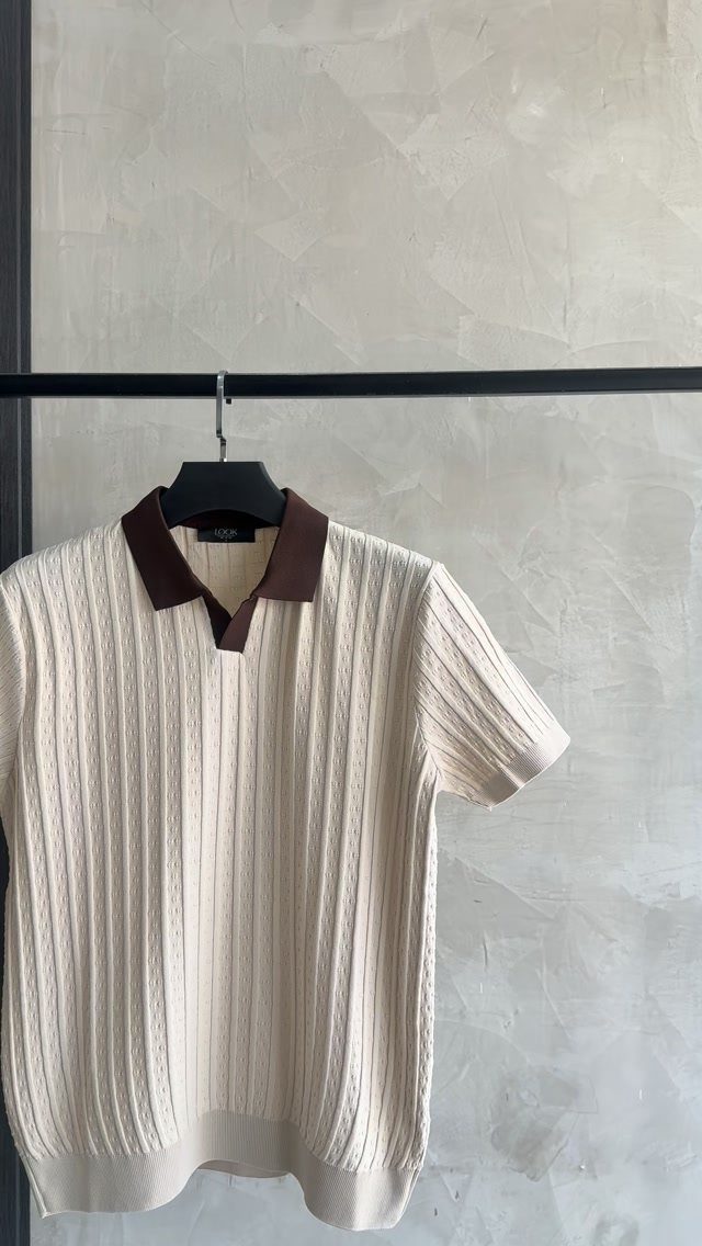A cream cable-knit polo shirt with a brown collar hanging on a black clothing rack.
