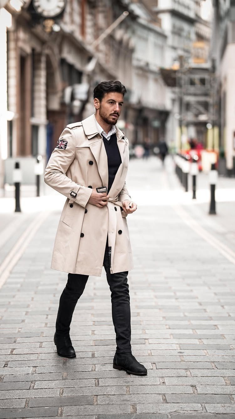 A man posing in a beige trench coat, dark sweater, and black trousers on a city street.