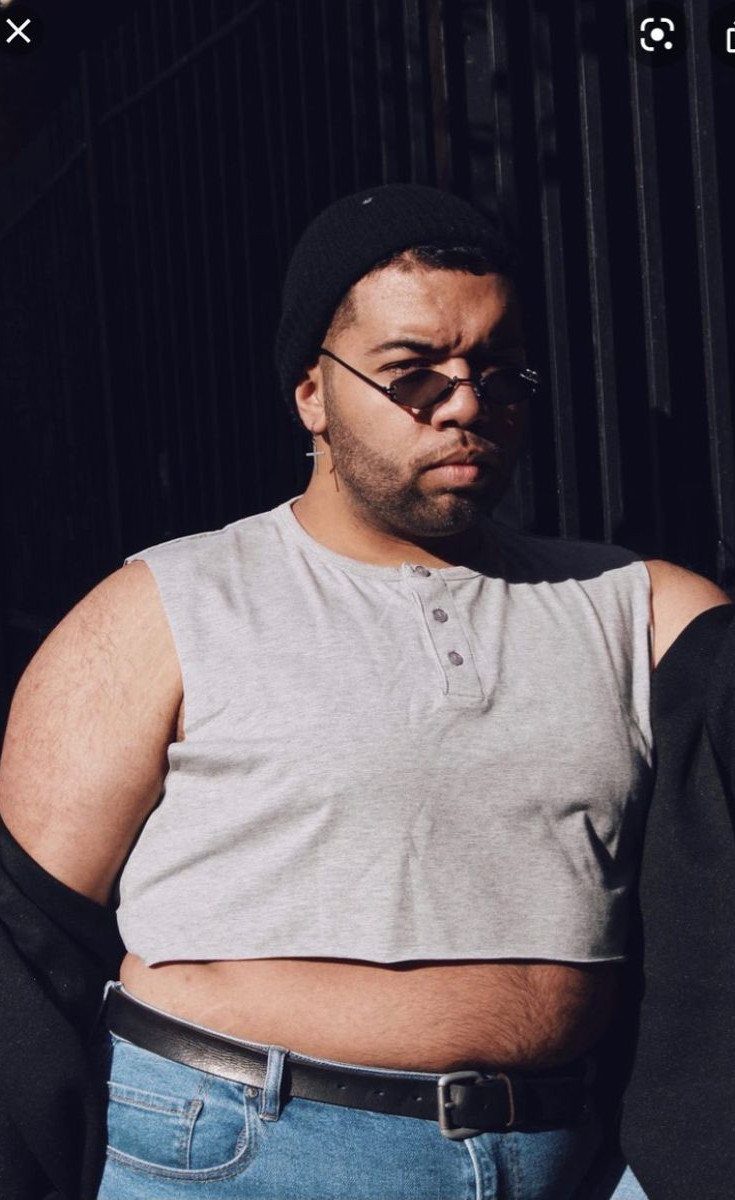 A man with a larger build wearing a grey crop top, sunglasses, and a black beanie.