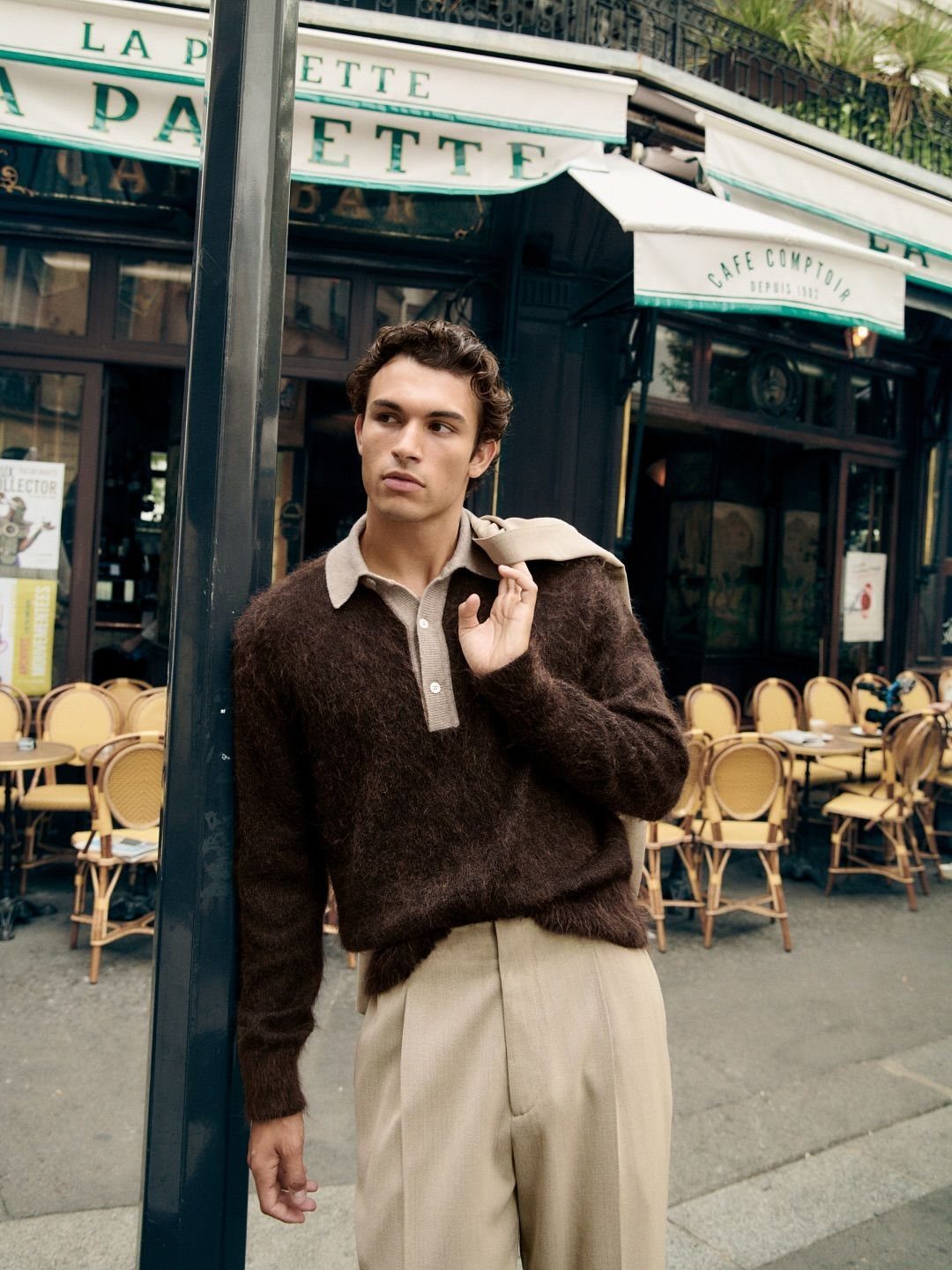 A male model posing in a brown sweater and beige trousers outside a Parisian cafe.