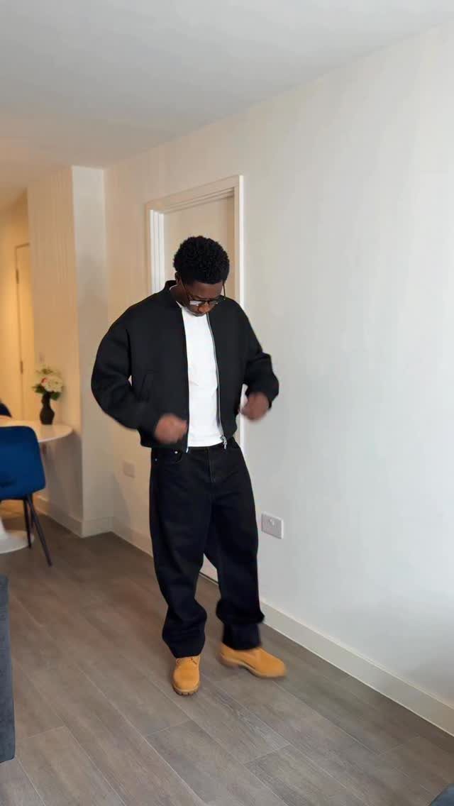 A man wearing a black bomber jacket, white t-shirt, black pants, and tan boots.