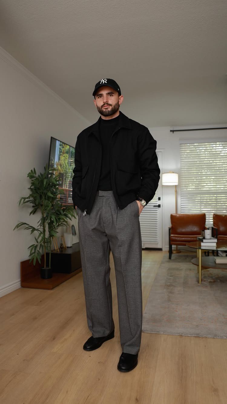 A man posing in a black jacket, black turtleneck, grey trousers, and a black baseball cap.
