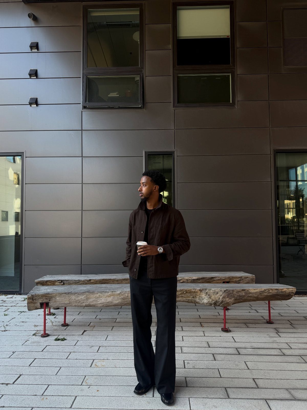 A man in a dark brown jacket and black trousers standing near a stone bench.