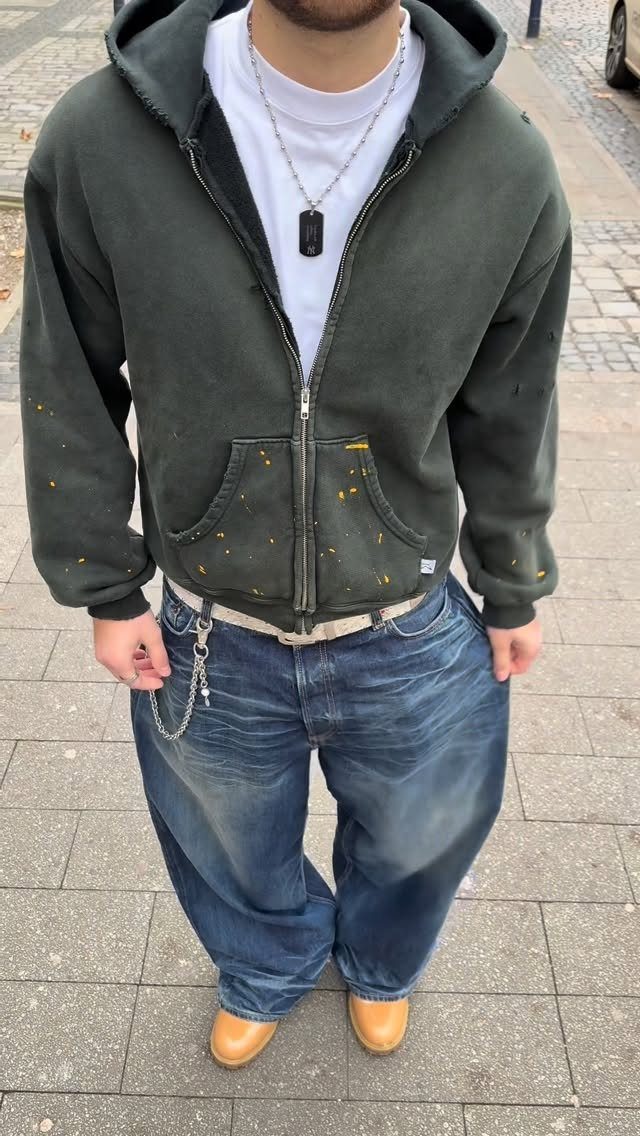Man wearing a dark green hoodie, wide blue jeans, and tan leather shoes standing on a sidewalk.