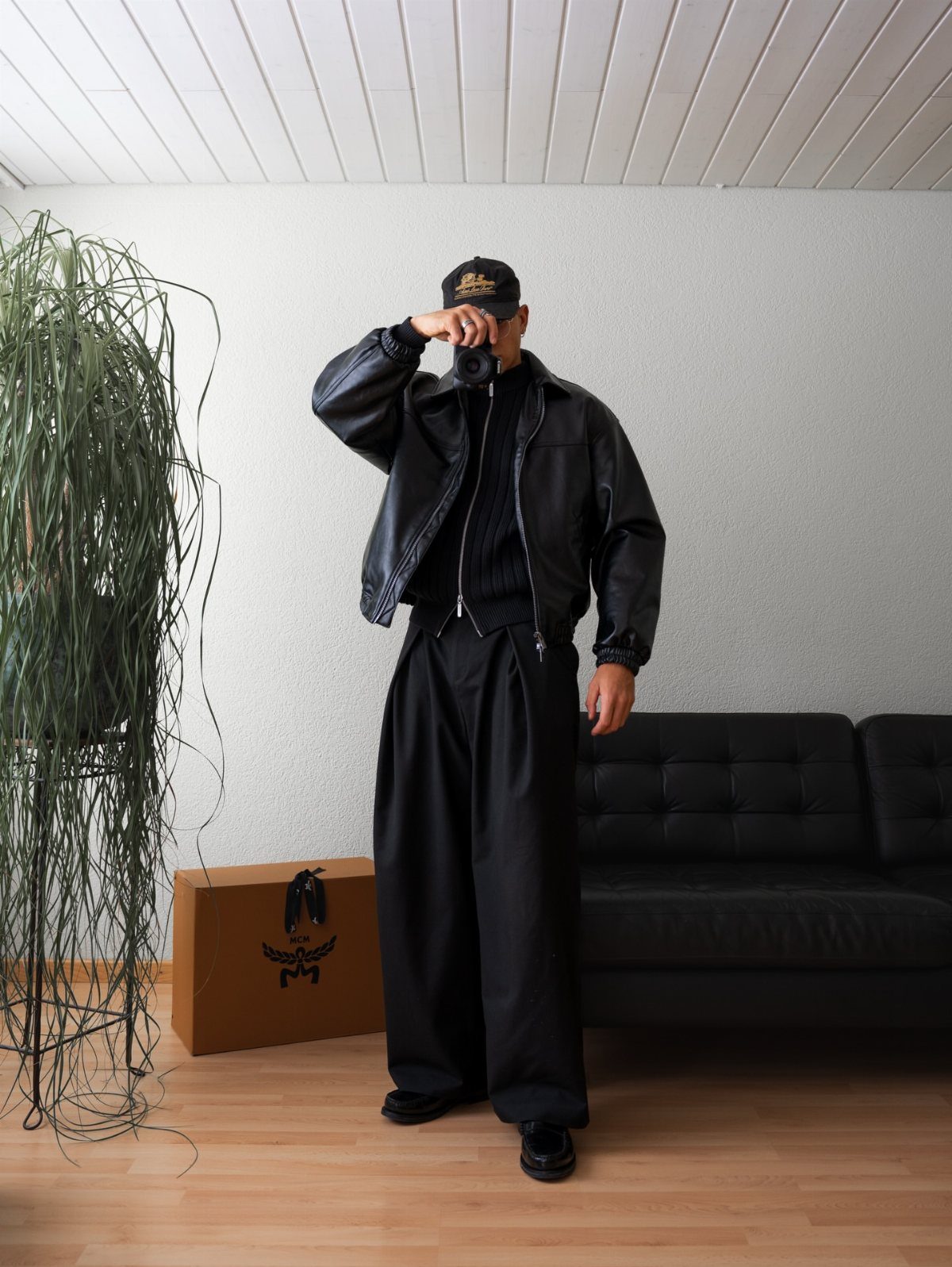 A man in an all-black outfit posing with a camera in a minimalist room.