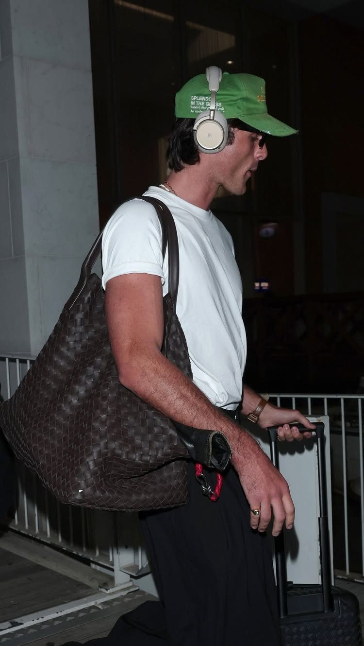 Man in a white t-shirt and green cap carrying a large brown bag and pulling a suitcase.